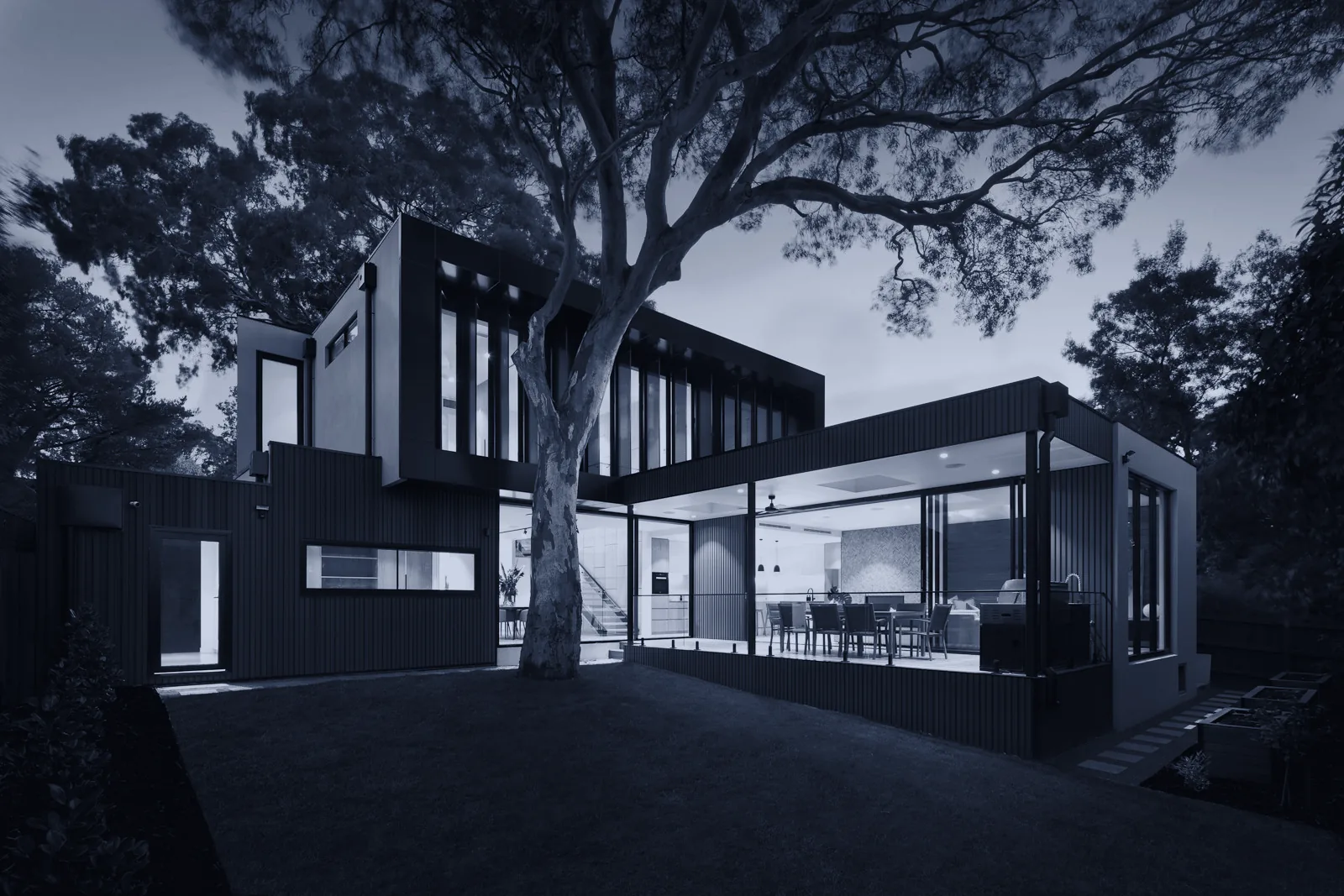 A modern house lit at dusk under a large tree